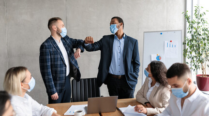 Two Businessmen In Face Masks Bumping Elbows During Corporate Meeting Standing In Modern Office. New Handshake Greeting, Etiquette For Business People During Coronavirus Pandemic