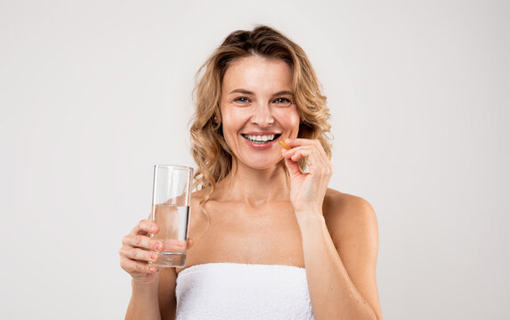 Beauty Supplement. Attractive Middle Aged Woman Holding Vitamin Capsule And Water Glass, Smiling Female Taking Pill For Beautiful Skin, Hair And Nails, Standing Wrapped In Towel On Light Background