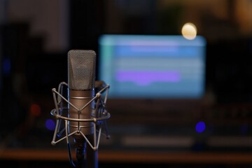 Studio condenser mic in foreground recording sound, with blurred computer screen displaying audio waveform in background