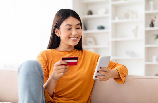 Fast Online Shopping. Smiling young chinese lady holding debit credit bank card and using cell phone, sitting on couch at home in living room, looking at smartphone screen, free copy space