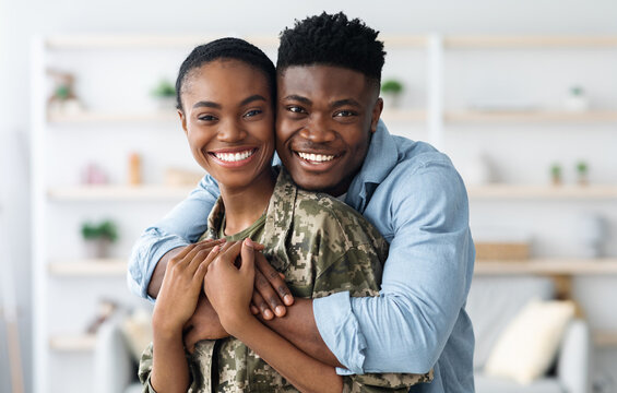 Closeup portrait of cheerful black female soldier lady in camouflage uniform hugging with boyfriend, home interior. Military wife came back from army, bonding with loving husband, copy space