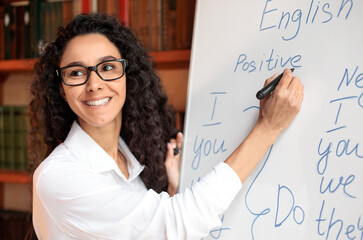 Lecture. Closeup portrait of cheerful smiling female tutor wearing glasses teaching English language, writing grammar rules on the board with marker, looking back at her students at classroom