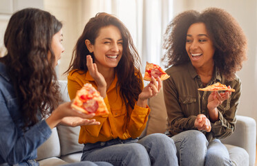 Three diverse women friends enjoying snacks and joyful chat, laughing together while eating pizza on a cozy sofa in modern living room interior. Home gathering, female friendship