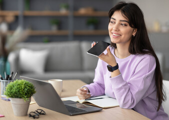 Portrait of smiling cheerful young woman sitting at desk using laptop, talking on speakerphone,...