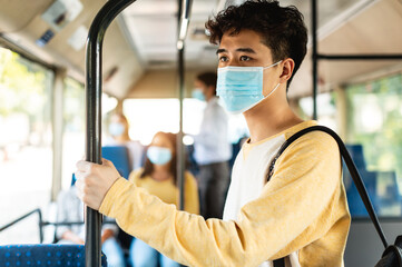 Travel Safely On Public Transport. Portrait of young asian man in protective face mask looking through train window. Male passenger with backpack riding standing inside autobus, holding handle