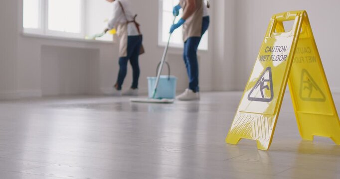 Yellow caution sign warns of a wet floor, while cleaning service staff mop the area in the background. The safety warning ensures caution and prevents slips during the cleaning process.