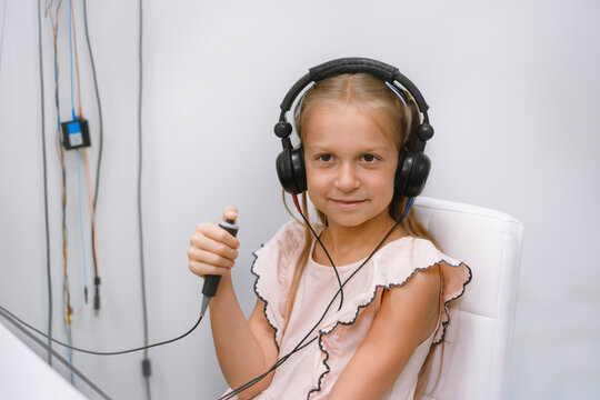 Smiling girl in audiometry booth wearing headphones and responding to tones during pediatric hearing test in medical clinic