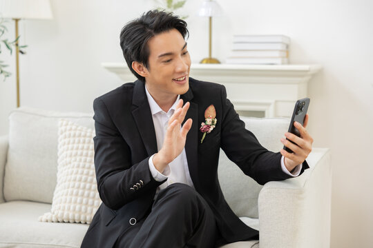Asian Young man in black suit sitting on sofa waving hand during video call on smartphone, modern living room interior, cheerful communication