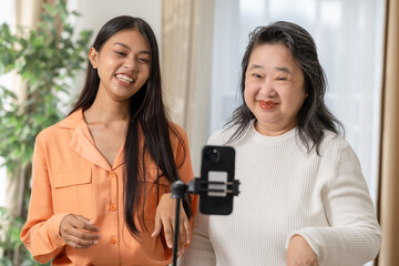 Asian Two women smiling and recording video together using smartphone on tripod in bright room with natural light and plants