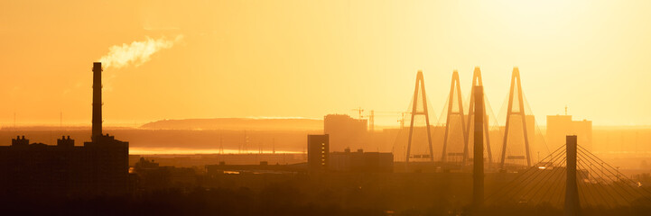 Cityscape of Saint Petersburg at sunrise with cable-stayed bridge, chimneys and buildings in warm golden light and morning haze.