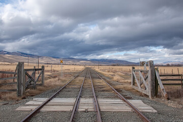 Rural railway crossing with wooden gates and dirt road leading across train tracks into open countryside under a bright blue sky with clouds.