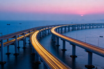 Obraz premium A long curved bridge over water at twilight with illuminated roadways and a pink sky