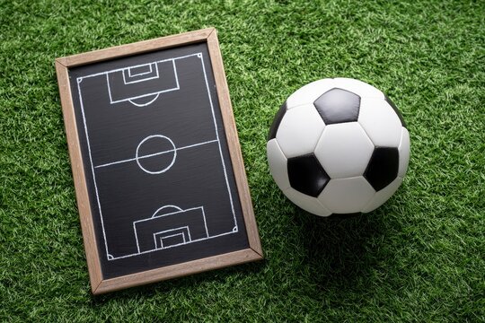 A soccer ball sits on green grass alongside a chalkboard diagram of a field presenting a sports setting - Powered by Adobe