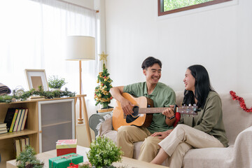 A cheerful couple enjoys the holiday season as the man plays guitar, the woman smiles beside him on the sofa, a christmas tree and wrapped gifts are nearby creating a festive atmosphere in their home