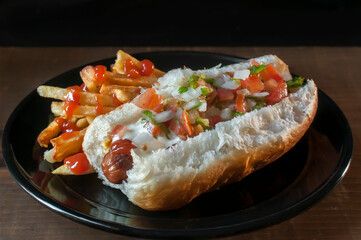 Hot dog with pico de gallo and cream on top, with fries on one side on a black plate and dark background