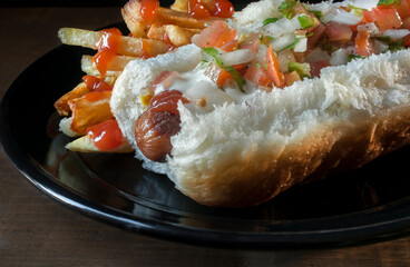 Hot dog with pico de gallo and cream on top, with fries on one side on a black plate and dark background, maro photpgraphy or close up