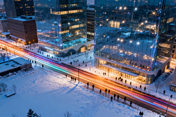 Aerial view of modern glass buildings in a snowy urban area at night with light trails from traffic and pedestrians