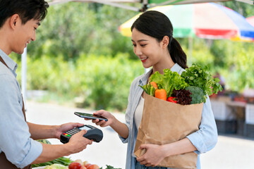 A woman purchasing fresh vegetables at an outdoor market using a contactless payment method.