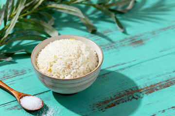 A bowl of cooked white rice on a teal wooden surface with a wooden spoon containing salt and green foliage in the background.