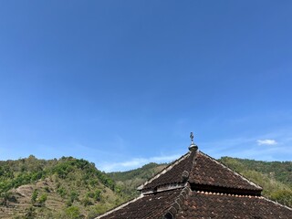 Traditional tiled roof of a rural mosque building with small dome beneath clear blue sky, with green hills in the background, highlighting countryside architecture and nature.