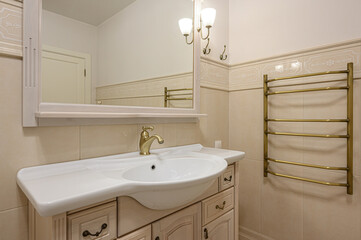 A bright, elegant bathroom with a white sink, vanity, gold faucet, mirror, and towel warmer. Beige tile adorns the walls