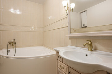 A well-lit bathroom features a white tub and sink with gold fixtures, framed by cream-colored tile. A mirror and wall-mounted lights add to the room's classic aesthetic