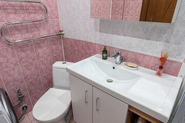 A cozy bathroom corner with pink floral tiles, a white sink, toilet, and heated towel rack. It has a homely, clean feel