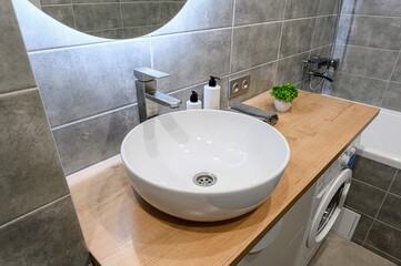Modern bathroom with a round, white sink on a wood counter. Grey tiles cover the walls. A silver faucet and toiletries are near the sink. A round mirror is above
