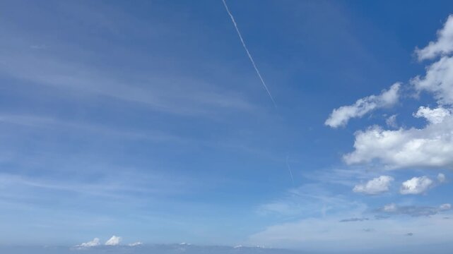 Blue sky with thin airplane contrail line and scattered white clouds in bright natural background
