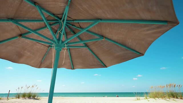 Low Angle Under Beach Umbrella View on Sandy Coastline with Blue Sky Above. Calm Ocean Scene with Distant Figures and Sparse Vegetation Near Turquoise Parasol Frame Creates Summer Ambiance