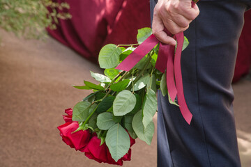 Close-up of an anonymous senior man in a suit holding a bouquet of red roses with ribbon; reserved male congratulations at a formal event. Photo