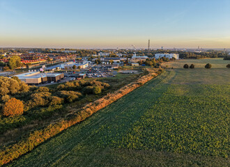 Aerial view of a town’s outskirts where industry meets open green fields, bathed in warm morning...