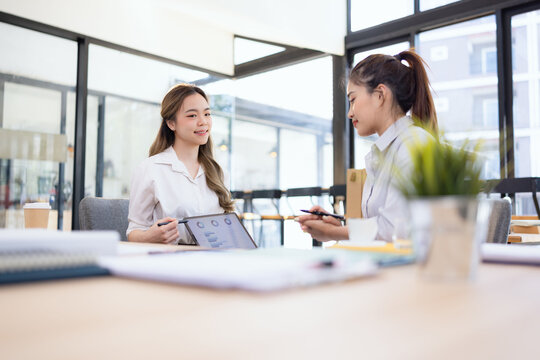 Two businesswomen discussing work using digital tablet and laptop in modern office, Business team analyzing financial data on a digital tablet and paper report.