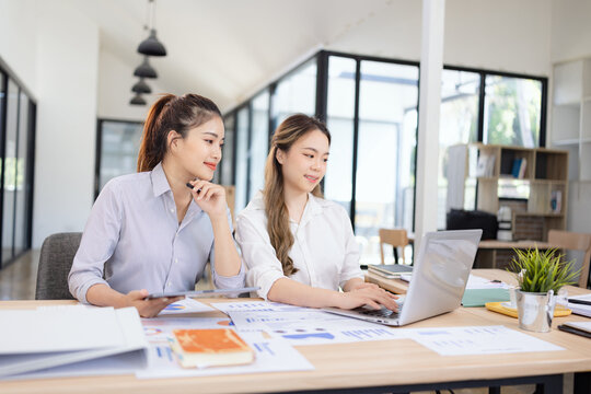 Two businesswomen discussing work using digital tablet and laptop in modern office, Business team analyzing financial data on a digital tablet and paper report.