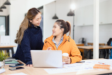 Two cheerful diverse office workers women cooperating on project, sitting at work desk with laptop, looking at screen, smiling.