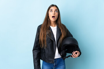 Young caucasian woman holding a motorcycle helmet isolated on blue background looking up and with surprised expression