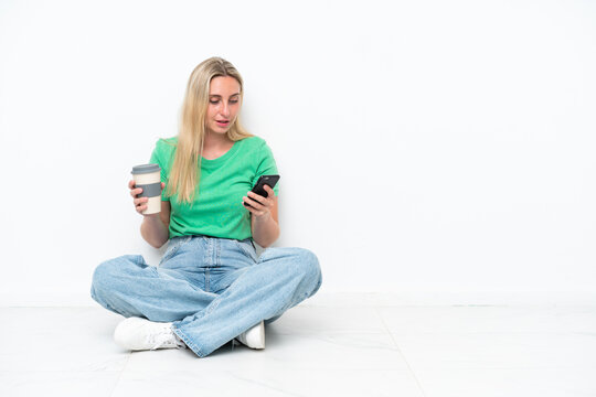 Young caucasian woman sitting on the floor isolated on white background holding coffee to take away and a mobile