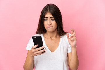 Fototapeta premium Young caucasian woman isolated on pink background using mobile phone with fingers crossing