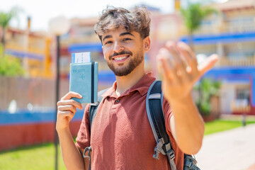 Young Arabian handsome man at outdoors holding a passport and doing coming gesture
