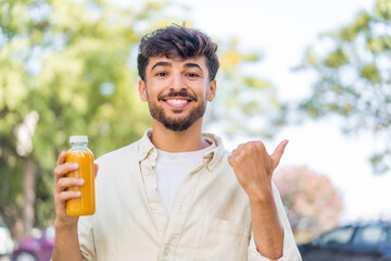 Young Arabian handsome man holding an orange juice at outdoors pointing to the side to present a product