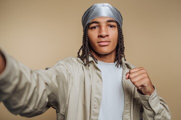 Young man with braided hair wearing a silver durag and beige shirt, posing confidently with a fist raised, against a neutral background.