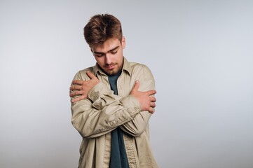 Young man hugging himself, wearing a beige shirt, against a plain background.