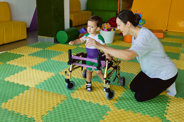 Handsome young boy with movement disorders practices walking with support from a therapist in rehabilitation clinic
