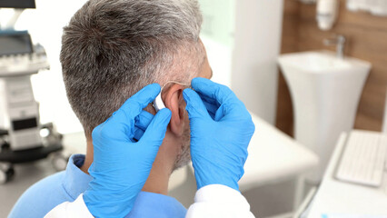 Fototapeta premium Audiologist installing hearing aid on ear of adult man in hearing clinic. Deafness Treatment, hearing solutions