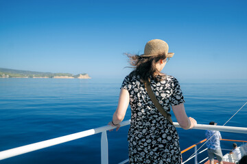 Rear view of a woman in a floral dress and straw hat standing on the deck of a ferry or tour boat in Greece. She’s looking at the calm blue Aegean Sea with an island coastline in the distance under 