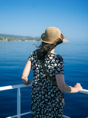 Rear view of a woman in a floral dress and straw hat standing on the deck of a ferry or tour boat in Greece. She’s looking at the calm blue Aegean Sea with an island coastline in the distance under 