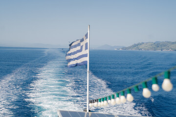 Greek flag waving on the back of a ferry as it sails across the clear blue waters of the Aegean Sea. Decorative string lights on the deck create a festive and relaxing holiday atmosphere.