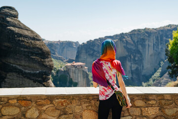 Back view of a woman wearing a vibrant, multicolored scarf while standing at a stone historic monasteries of Meteora, Greece. The photograph captures a serene moment of travel and cultural exploration