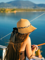 A young woman with long hair and a straw hat standing on the deck of a boat, looking out at the calm blue Aegean Sea and surrounding Greek islands.