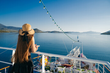 A young woman with long hair and a straw hat standing on the deck of a boat, looking out at the calm blue Aegean Sea and surrounding Greek islands.